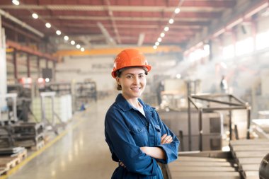 Mutlu younh iş kıyafeti ve kask içinde fabrika başarılı kadın işçi kamera önünde dururken göğüs kollarını geçerken