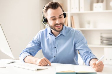 Kulaklık masa başında oturan, müşterilere danışmanlık ve not defterinde çalışma notları yapma ile mutlu genç adam