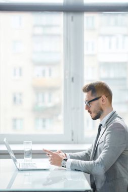 Young serious office manager in formalwear using modern gadgets while sitting by desk