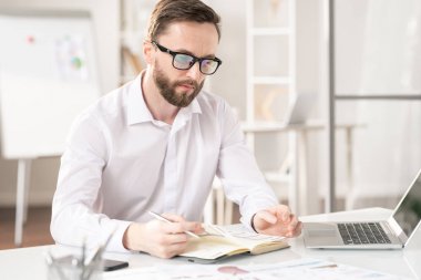 Young elegant professional making notes in notebook while writing down working plan or agenda in the morning