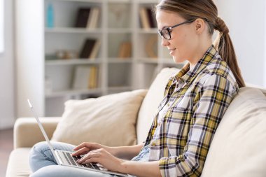 Online dosyaları ile çalışırken rahat kanepede oturan ve dizüstü bilgisayarda Internet kullanarak gözlük pozitif çekici genç kadın
