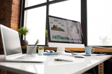Ekran, dizüstü bilgisayar, kupa ve diğer malzemeleri ofis penceresi ile masa üzerinde grafik bilgileri ile bilgisayar monitörü