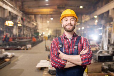 Young smiling professional in overalls and protective helmet standing in front of camera inside large machinebuilding plant