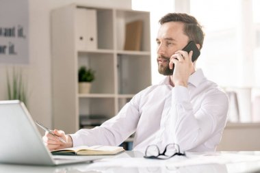 Young man with smartphone by his ear looking at laptop display while calling one of clients by workplace