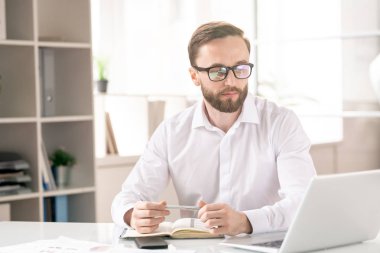 Young bearded businessman in eyeglasses and white shirt looking at laptop display while organizing work in office
