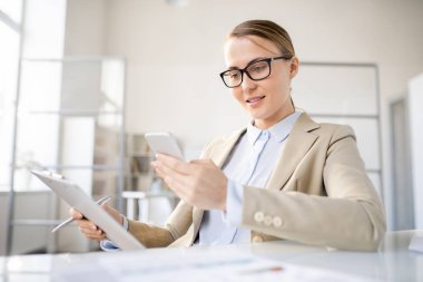 Bej ceketli gülümseyen genç iş kadını masada otururken ve belgeanaliz ederken akıllı telefondaki mesajı kontrol