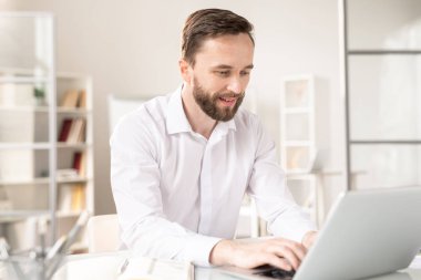 Young analyst in white shirt typing on laptop keypad while netering information or browsing in the net