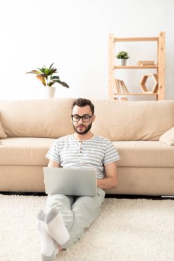 Young relaxed businessman or freelancer in casualwear sitting on the floor by couch while carrying out remote work at home