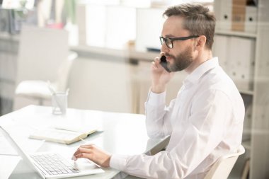 Young handsome bearded businessman in eyeglasses talking by smartphone with client while scrolling in the net in front of laptop