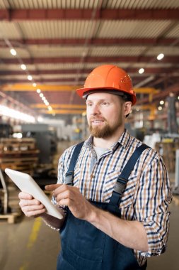 Young factory technician scrolling through online data in touchpad while controling working process