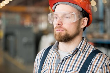 Modern fabrika veya sanayi fabrikasının sakallı genç mühendisi kamera önünde atölyede duruyor