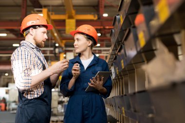 Young male and female engineers in helmets and overalls discussing quality of new technical supplies in workshop