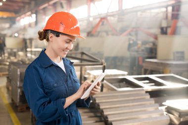 Young successful worker of contemporary factory in workwear and helmet scrolling in touchpad while searching for technical data