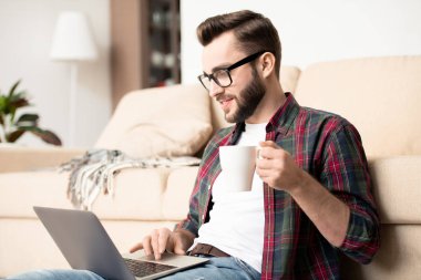Young casual employee with mug of tea and laptop sitting by couch in living-room and working remotedly
