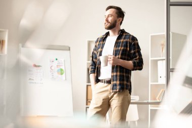 Young restful man with mug standing in his office and enjoying tea break in the middle of working day
