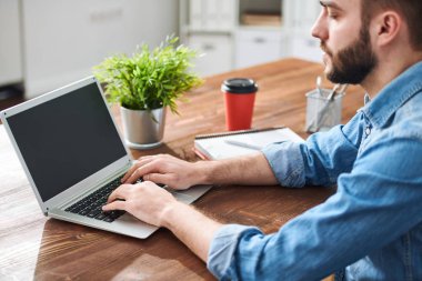 Young casual businessman typing on laptop keypad by table while entering information in database or planning work