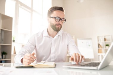 Young office manager or analyst concentrating on work with online data while sitting by desk in front of laptop