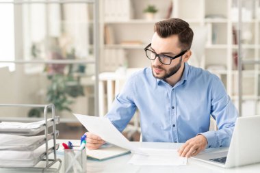 Young serious businessman in eyeglasses reading financial paper or contract while sitting by desk in office
