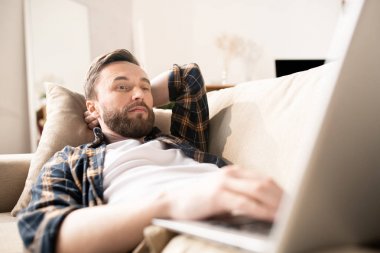 Young pensive bearded man in casualwear lying on couch with laptop in front and watching online video or reading information in the net
