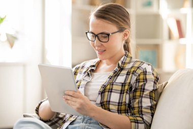 Online kursa katılırken rahat kanepede oturan ve dijital tablet kullanan gözlüklü neşeli akıllı kız