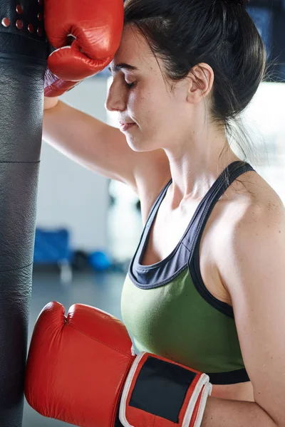 Young tired or stressed woman in boxing gloves standing by punch bag during workout in gym