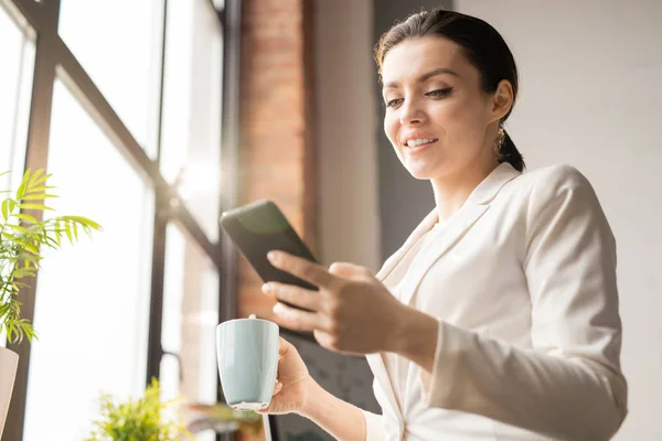 お茶を飲みながら オフィスの休憩時にスマートフォンでフォーマルウェアメッセージングで成功した若いビジネスウーマン — ストック写真