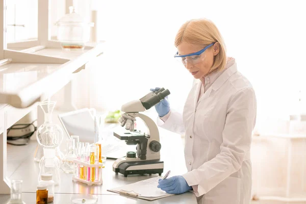 严重集中的女士生物学家在实验室外套和安全护目镜使用显微镜和观察观察在实验室 — 图库照片