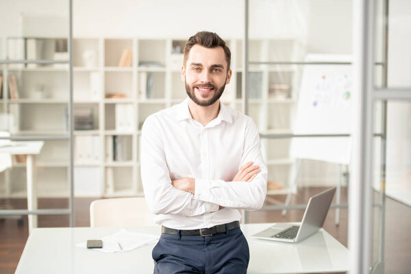 Young elegant office manager or broker crossing his arms on chest while standing by workplace in front of camera