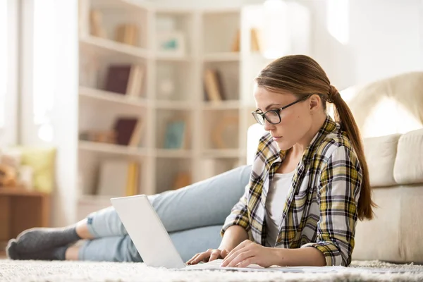 自宅で仕事をしながら カーペットの上に横たわってノートパソコンを使用してポニーテールを持つ深刻なペンシブ若い女性フリーランサー — ストック写真