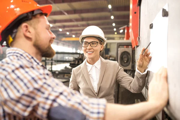 快乐的年轻女工程师在头盔指着白板上的纸张 同时为同事做演示 — 图库照片