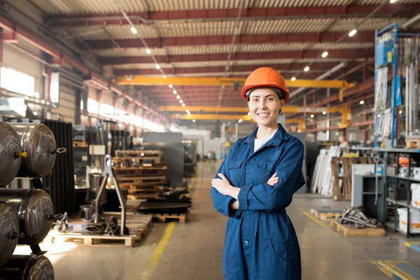 快乐的年轻女工的大型现代工厂的蓝色制服和防护头盔交叉她的手臂在胸前 — 图库照片