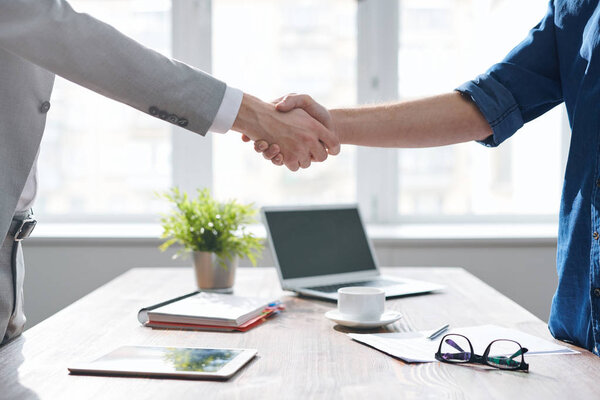 Handshake of two young business partners over desk with office supplies after signing contract at meeting