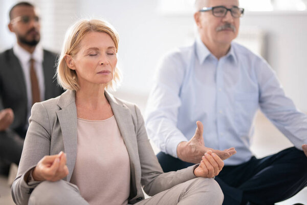 Пожилая блондинка в костюме и ее коллеги медитируют во время перерыва, сидя в позе лотоса
