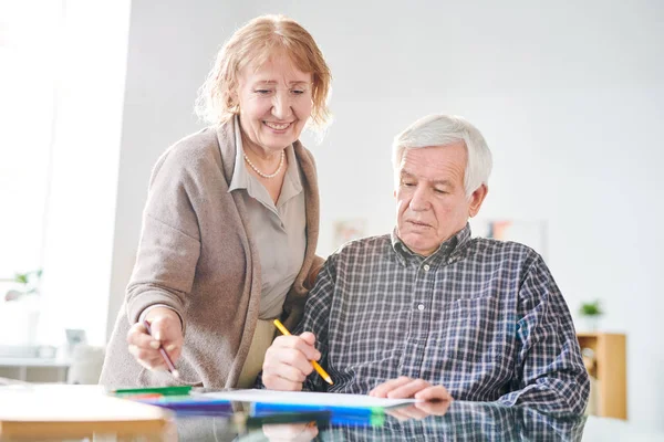Senior handicapped man and happy aged casual woman discussing creative ideas while drawing together with crayons in retirement home