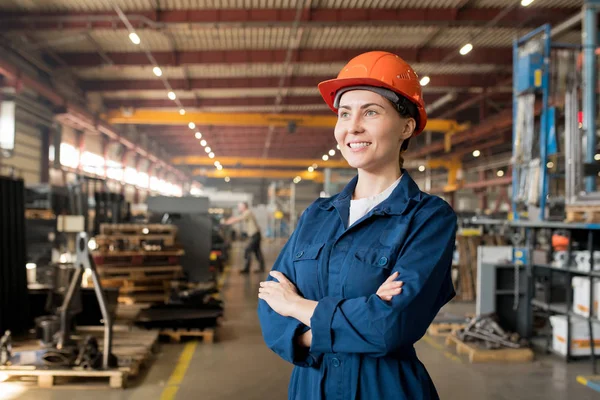 穿着蓝色工作服和头盔的快乐年轻女技师站在工厂大车间内 — 图库照片