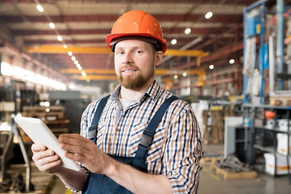 Young male engineer in helmet and overalls looking for technical data in the net while working in factory