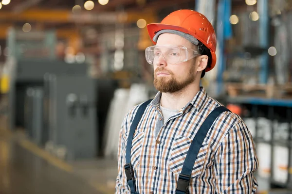 在现代化的大工厂里 戴着头盔 眼镜和工作服的年轻而严肃的工程师站在摄像机前 — 图库照片