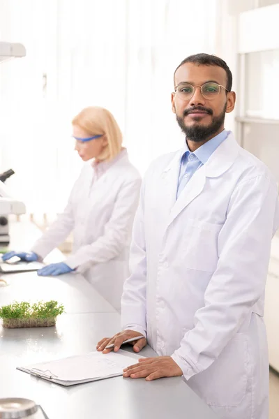 Portrait of content confident young middle-eastern laboratory scientist in white coat standing at lab bench and making notes in paper