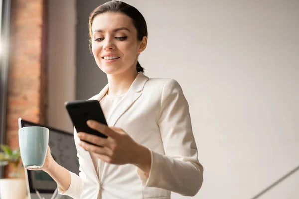 オフィスのコーヒーブレイク時にスマートフォンでメッセージや通知を読み取る白いスーツで成功した女性オフィスマネージャー — ストック写真