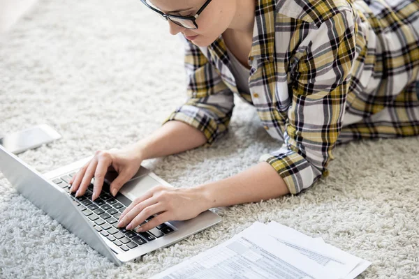 ラップトップ 納税申告の概念にデータを入力しながら カーペットの上に横たわってキーボードにタイピングメガネで深刻な忙しい若い女性 — ストック写真