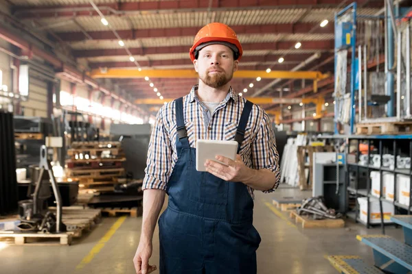 在一家大型工厂的车间里 身穿工作服头戴头盔 留着胡子的年轻人站在摄像机前 — 图库照片