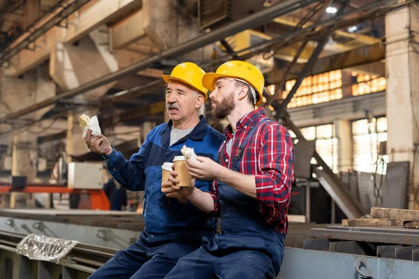 Two restful technicians with drinks and snacks sitting in workshop and discussing new equipment at lunch break