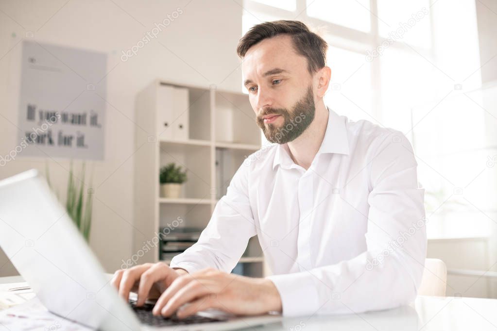 Bearded analyst or broker in white shirt looknig at laptop display while browsing in the net and searching for online data