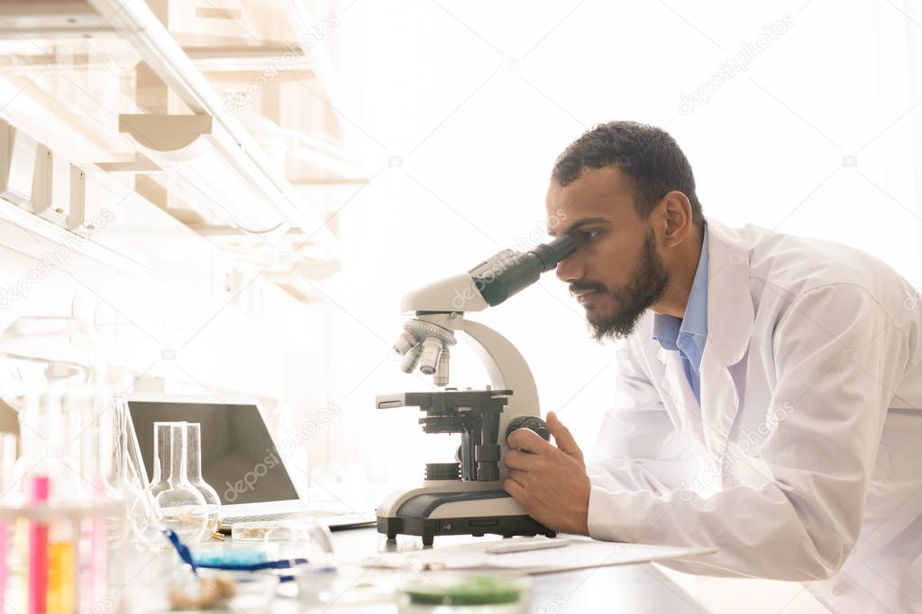 Concentrated young Arabian scientist with beard and mustache standing at desk and using modern microscope in laboratory