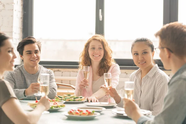 陽気な女の子とシャンパンを持つ男は ホームパーティーで提供されたテーブルでトーストを発音する彼らの友人を見て — ストック写真