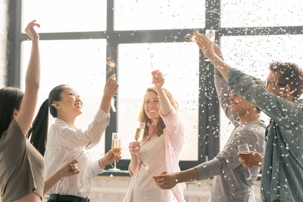 ホームパーティーで紙吹雪で踊りながらシャンパンで乾杯ベンガルライトで興奮した異文化の友人 — ストック写真