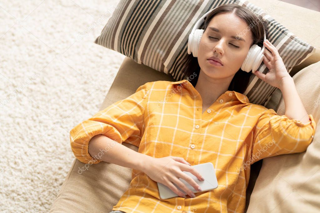 Young serene brunette female in headphones lying on couch while listening to relaxing or her favorite music at leisure