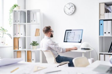 Genç çağdaş mühendis bilgisayar monitörünün önünde oturuyor ve inşaatın elektronik taslaklarına bakıyor.