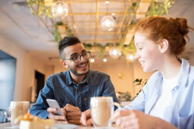Kafede otururken onun akıllı telefon onun kız arkadaşı yeni uygulama gösteren gözlük ve rahat kot gömlek Mutlu adam