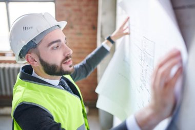 Şapkalı ve üniformalı genç bir mühendis tamamlanmamış binanın önünde dururken taslağa bakıyor.